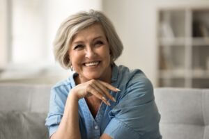 Smiling older woman with short gray hair and a light blue button-up shirt sits on a couch, resting her chin on her hand. The background is softly focused, showing shelves and a bright window.
