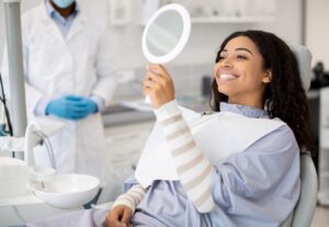 A woman sits in a dental chair, smiling and holding a round mirror. She wears a dental bib and long-sleeve shirt. A dentist in a white coat and blue gloves stands nearby, with dental tools in the foreground.
