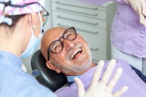 An older man with glasses smiles joyfully while reclining in a dental chair, wearing a bib. A dentist in a mask and gloves stands nearby, and another dental assistant is visible in the background.