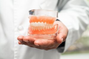 A person in a white lab coat holds realistic full upper and lower dentures encased in clear plastic in their right hand, displaying teeth and gums in a dental or medical setting.