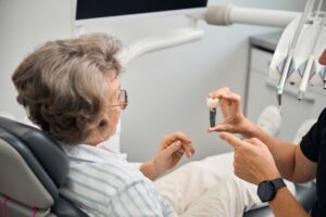 An older woman sits in a dental chair, looking at a dentist who is holding and explaining a dental implant model. The scene is set in a modern dental office with visible dental equipment.